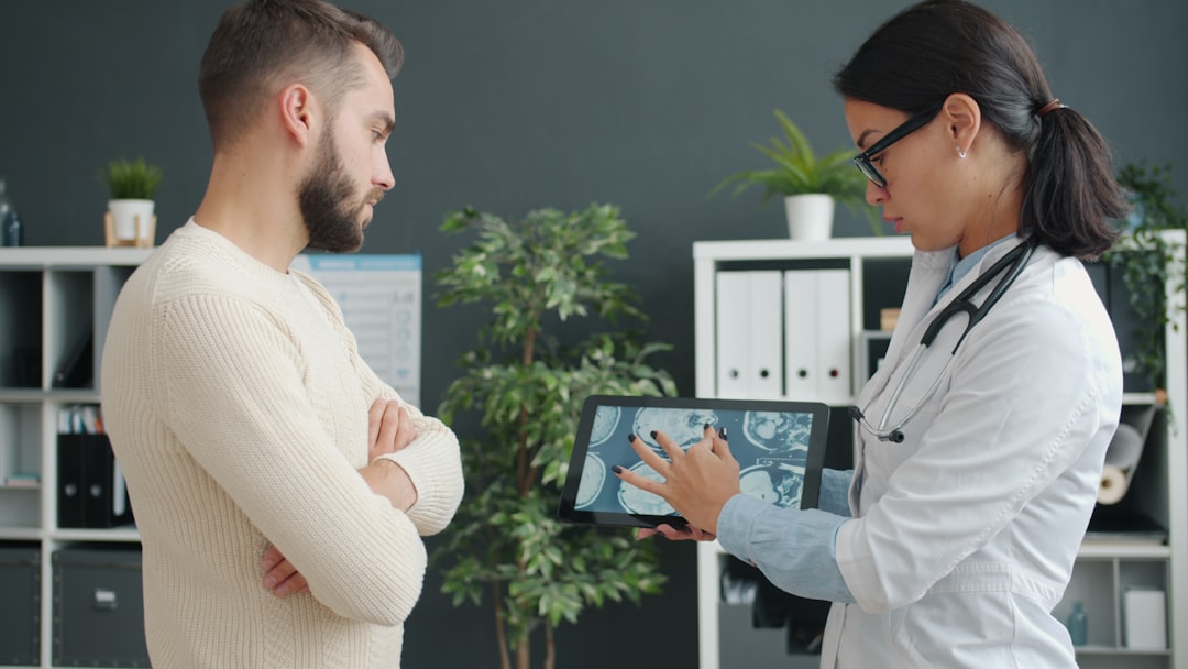 Female practitioner in uniform is showing male patient young man mri results on tablet screen in hospital office. Medical care and people concept.